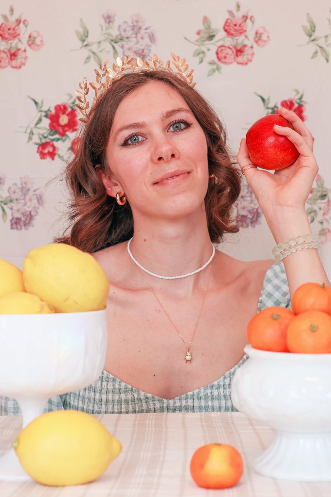 A girl sits at a table with a floral backdrop behind her, and bowls of fruit in front of her. She holds an apple.