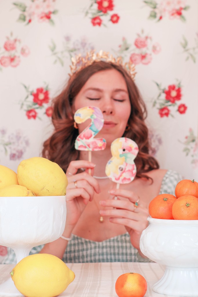 A girl sits at a table with a floral backdrop behind her, and bowls of fruit in front of her. She holds up the number "28".