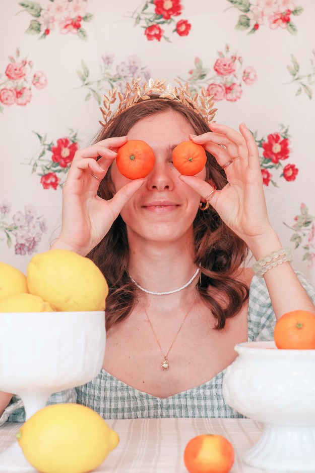A girl sits at a table with a floral backdrop behind her, and bowls of fruit in front of her. She holds two oranges up to her eyes.