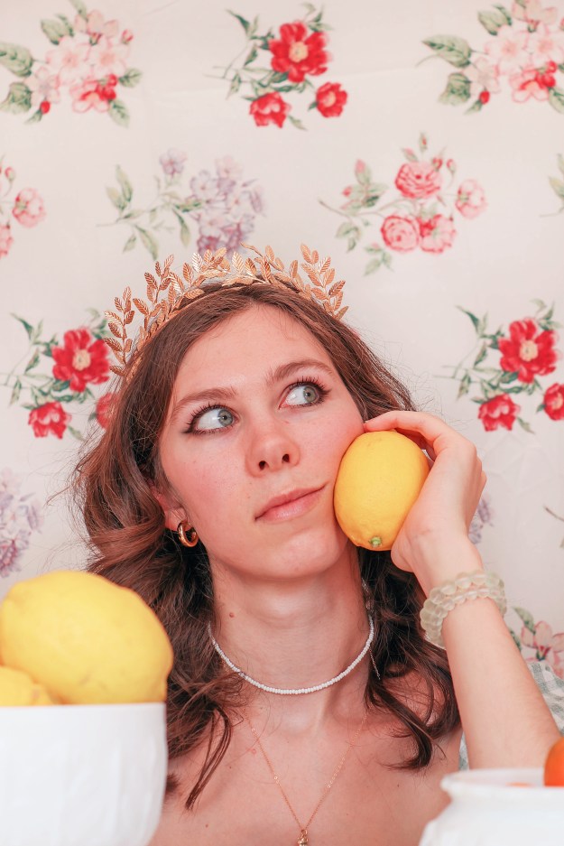 A girl holds a lemon up to her face in front of a floral backdrop.