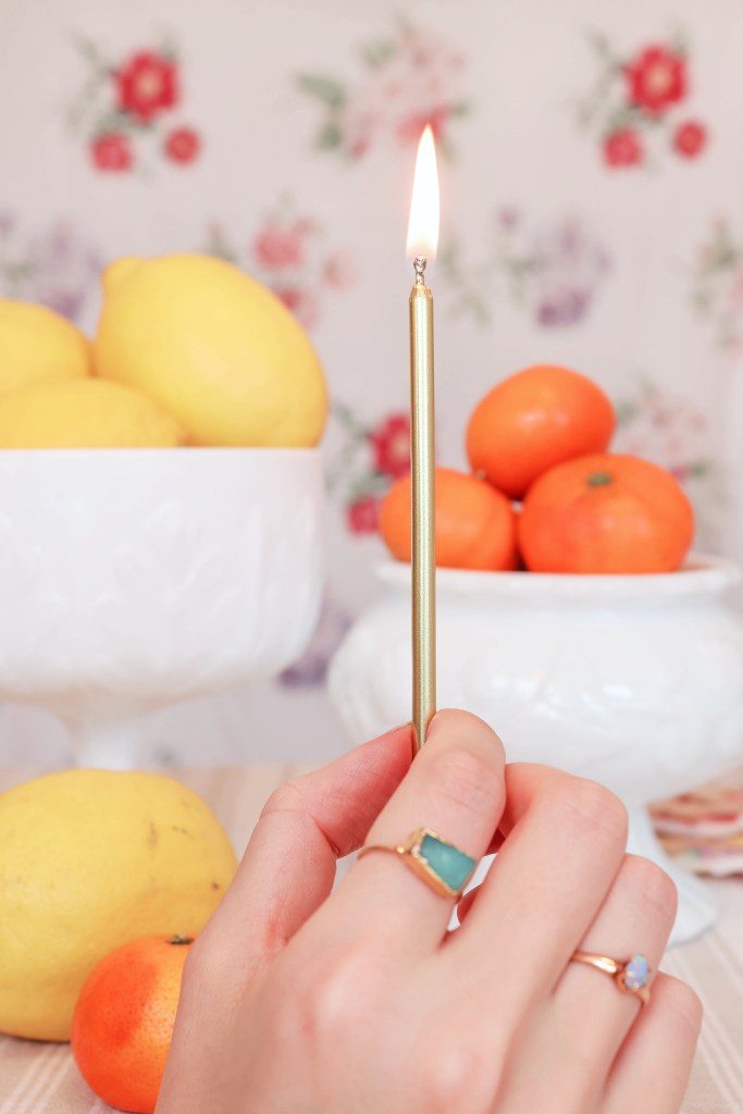 A hand holds a birthday candle in front of bowls of fruit.