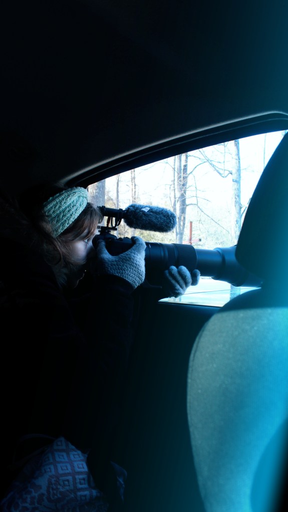 A girl holding a large camera looks out of a car window.