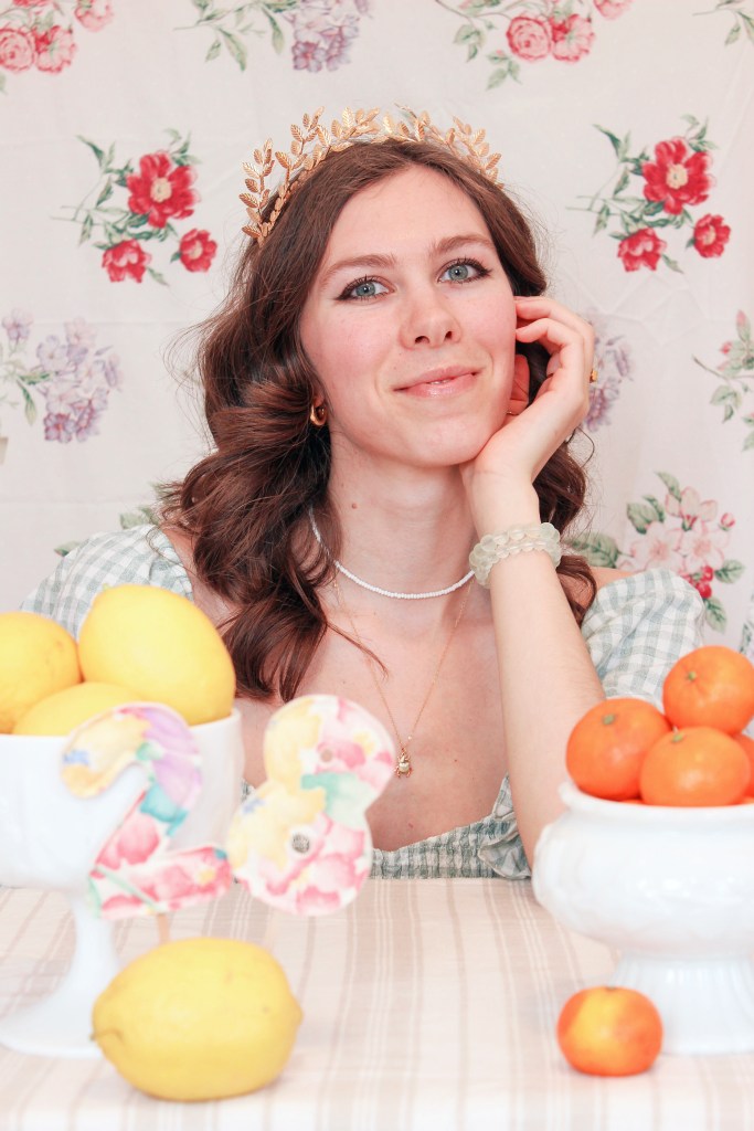 A girl sits at a table with a floral backdrop behind her, and bowls of fruit in front of her.