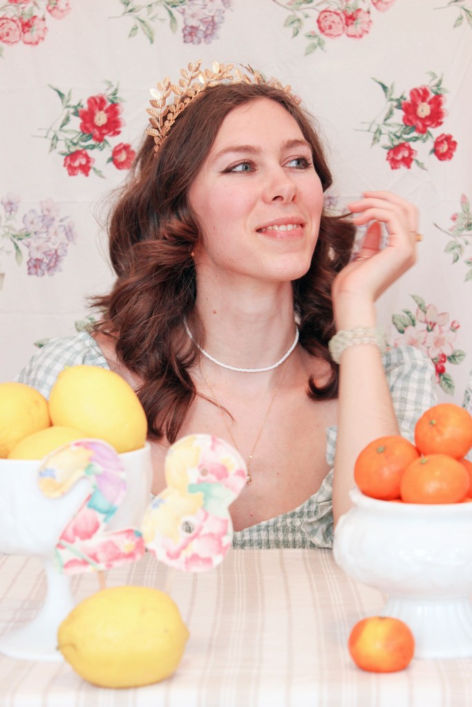 A girl sits at a table with a floral backdrop behind her, and bowls of fruit in front of her.
