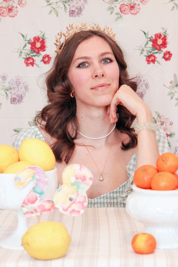 A girl sits at a table with a floral backdrop behind her, and bowls of fruit in front of her. 