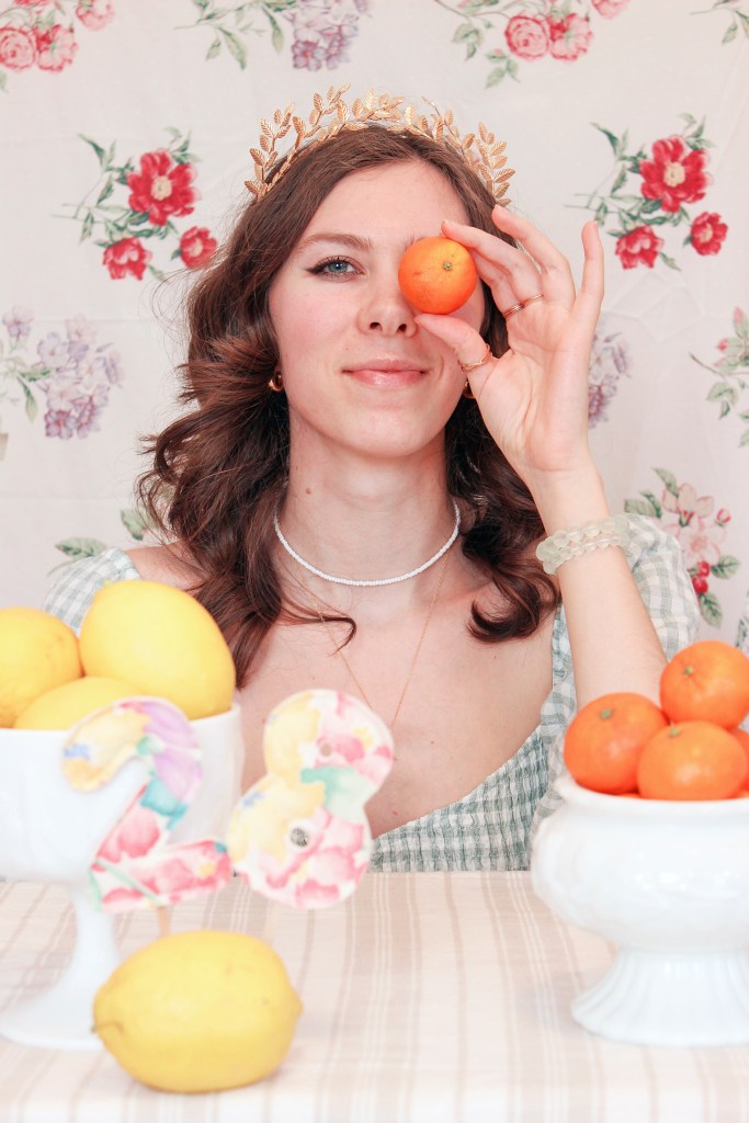 A girl sits at a table with a floral backdrop behind her, and bowls of fruit in front of her. She holds an orange up to her face.