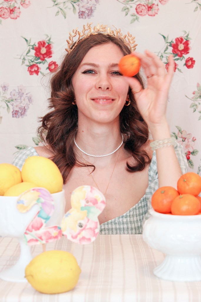 A girl sits at a table with a floral backdrop behind her, and bowls of fruit in front of her. She holds an orange up to her face.