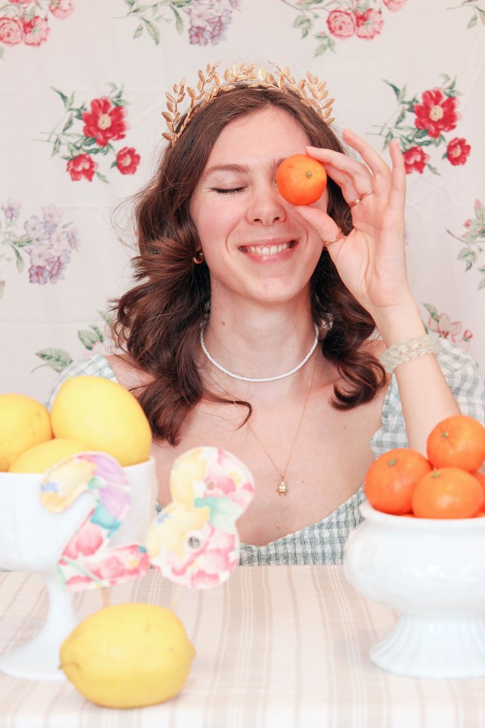 A girl sits at a table with a floral backdrop behind her, and bowls of fruit in front of her. She holds an orange up to her face.