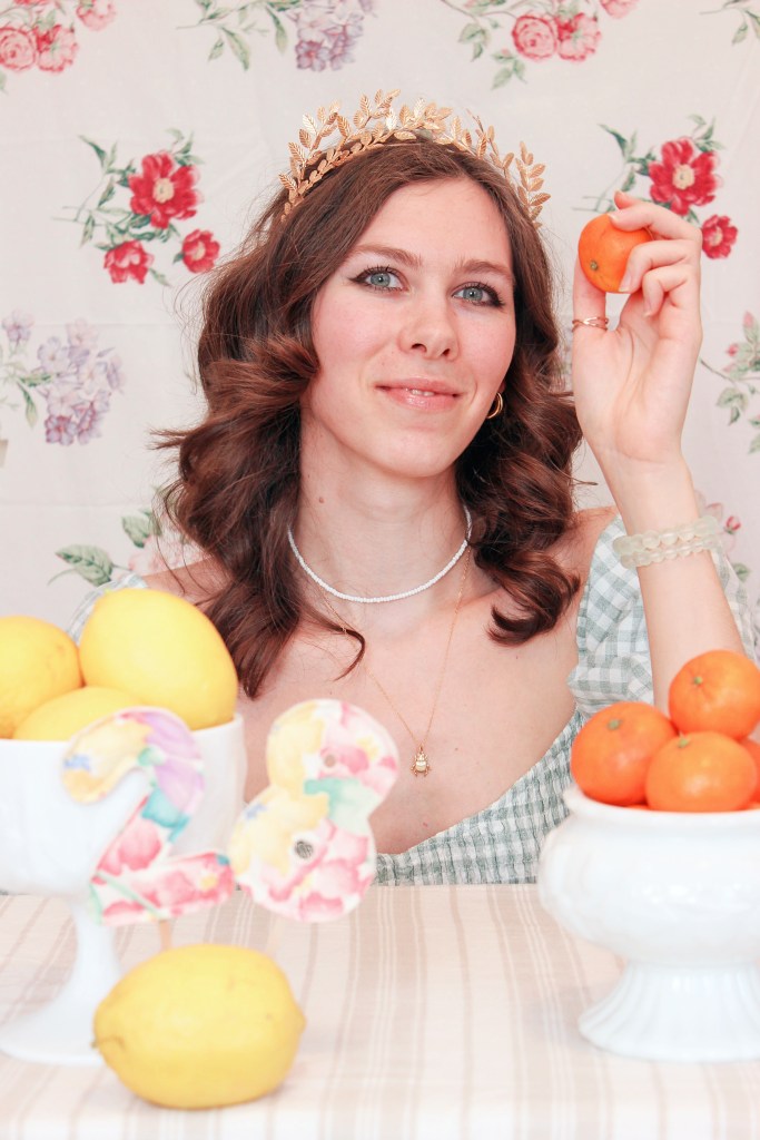 A girl sits at a table with a floral backdrop behind her, and bowls of fruit in front of her. She holds an orange up to her face.