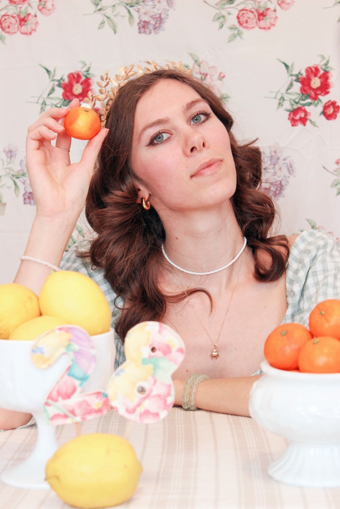 A girl sits at a table with a floral backdrop behind her, and bowls of fruit in front of her. She holds an orange up to her face.