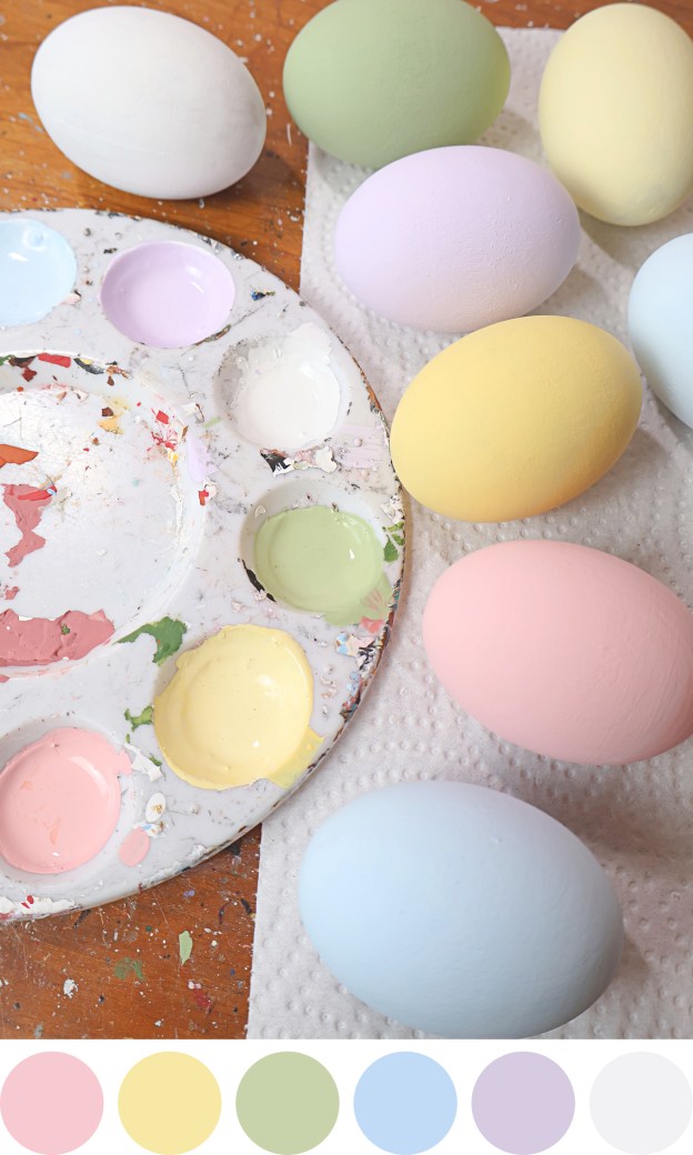 Wooden eggs in various pastel colors sit on a desk with a paint palette