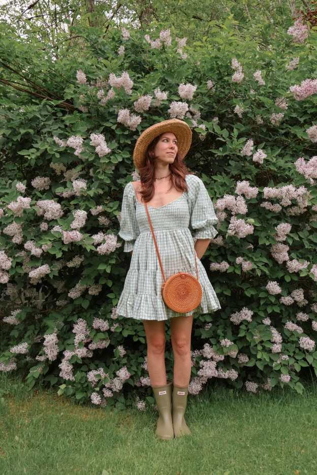 A girl wearing a checkered dress and straw sunhat stands in front of a big blooming lilac bush