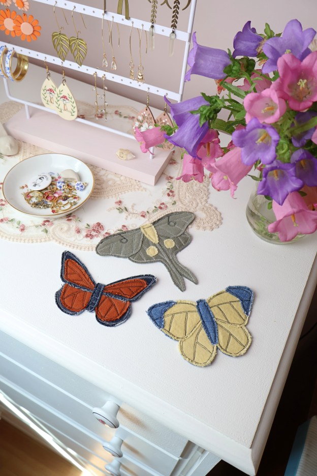 Three denim patches sewn into the shape of various butterflies and moths sit on a white dresser with a vase of flowers beside them.