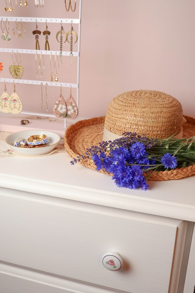 A straw hat with blue flowers laying on it sits aesthetically on a white dresser.