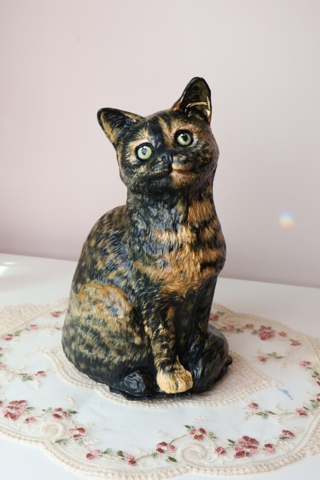 A statue painted to look like a tortoiseshell cat sits on a lace doily on a white dresser.
